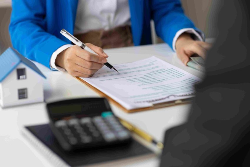 Person signing real estate paperwork at a desk with a calculator and model house nearby