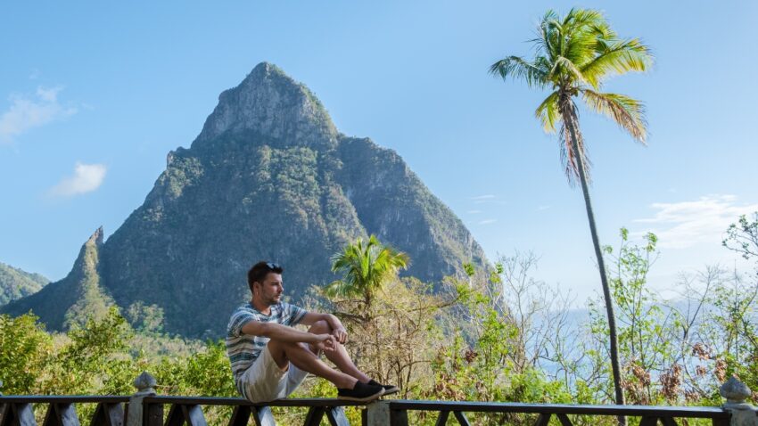 A man sits by a balcony rail with the Pitons and palm trees behind him in St. Lucia