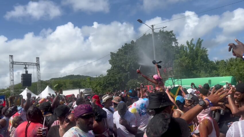 A crowd at St. Lucia Carnival sprays water near a stage under a blue sky